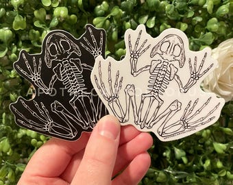 Op de afbeelding: Twee stickers van een skelet van een kikker, een zwarte en een witte. De witte sticker heeft de tekst "APOTHECARYOCTOPUS" onderaan.