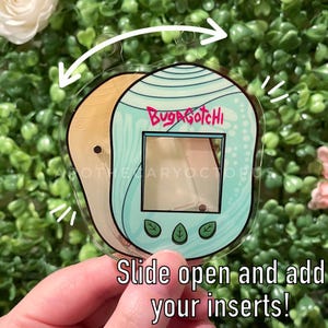 Puede incluir: Un llavero de plástico transparente con forma de Tamagotchi azul y blanco con el texto "Bug-a-Gotchi" en la parte delantera. El llavero está abierto y listo para que agregues tus propios insertos.