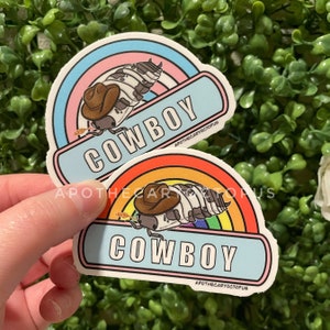 Puede incluir: Dos pegatinas con un bicho bola de dibujos animados que lleva un sombrero de vaquero y monta un arcoíris. El texto "COWBOY" está impreso en cada pegatina.