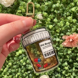 May include: A clear acrylic keychain shaped like a pill bottle, with the text "Pill Bugs" and "TAKE AS NEEDED". The bottle contains colorful pill bug illustrations. The keychain has a gold-colored clasp.