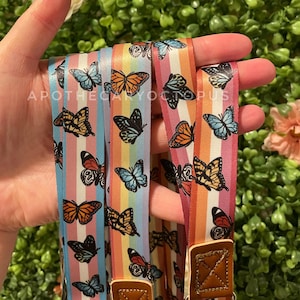 Puede incluir: Tres correas de cámara a rayas de colores con diseños de mariposas. Las correas son rosa, azul y naranja con rayas arcoíris. Cada correa tiene una correa ajustable de cuero marrón con un cierre metálico.