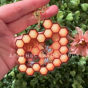 Könnte beinhalten: Ein Schlüsselanhänger in Form einer Honigwabe mit drei Bienen und einem Honigtropfen-Design. Der Schlüsselanhänger ist aus klarem Acryl mit einem roten und orangefarbenen Wabenmuster.