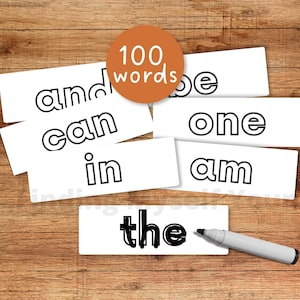 Op de afbeelding: Een verzameling witte rechthoekige kaarten met zwart omlijnde woorden zoals and, be, can, one, in, am en het woord the ingevuld met zwarte stift. Een grijze viltstift ligt naast de kaart met het woord the. Een oranje cirkel toont de tekst 100 words.