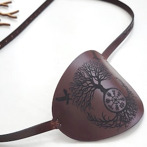 May include: A brown leather eye patch featuring a black laser-engraved design of a tree with a compass symbol integrated into its trunk and a bird silhouette. The patch is attached to a thin, adjustable brown leather strap with a metal buckle.