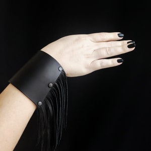 Könnte beinhalten: Ein schwarzes Lederarmband mit Fransen. Das Armband hat drei silberne Nieten.