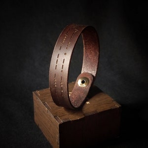 Può includere: Un braccialetto in pelle marrone con chiusura a bottone in ottone. Il braccialetto ha un motivo di punti e trattini incisi nella pelle.