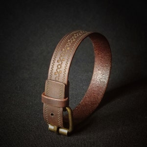 May include: A brown leather bracelet with a geometric pattern engraved on the surface. The bracelet has a brass buckle.