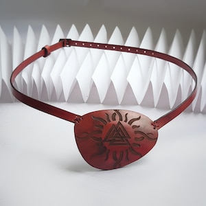 May include: A red leather eye patch with a sun and a Norse symbol, the Vegvisir, carved into it. The eye patch is attached to a brown leather strap.