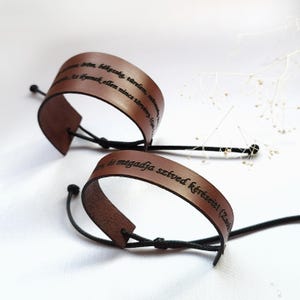 May include: Two brown leather bracelets with black cords. The bracelets have engraved text in a cursive font. The text on the top bracelet reads "mbékesség, türelem, te ilyenek ellen nincs törvény. (Ga". The text on the bottom bracelet reads "is megadja szíved kéréseiti (Zack".