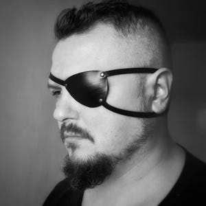 Puede incluir: Parche de ojo de cuero negro con correas ajustables. El parche para el ojo está asegurado con remaches y diseñado para cubrir un ojo. La imagen es un primer plano, que muestra el parche para el ojo en detalle.