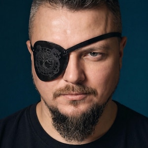 May include: Black leather eye patch with a decorative embossed design, worn over one eye. The eye patch is secured with a black strap. The person in the image is wearing a black t-shirt and has a beard.
