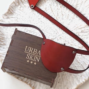 May include: A red leather eye patch with silver-toned metal rivets and a matching adjustable strap system. The eye patch is positioned next to a small, dark brown wooden box featuring the gold-colored text "URBA SKIN" and three stars.