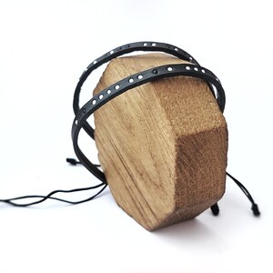 May include: Two black leather bracelets with white dots, tied together with a black cord, displayed on a wooden stand.