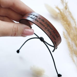 Puede incluir: Pulsera de cuero marrón con texto negro: «El que cree en mí, como dice la Escritura, de su interior correrán ríos de agua viva, Juan 7:38». La pulsera tiene un cordón negro ajustable.