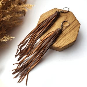 May include: A pair of long, brown leather tassel earrings. The earrings have a dark brown top with a loop and clasp, and the tassels are made of thin strips of lighter brown leather. The earrings are displayed on a wooden block.