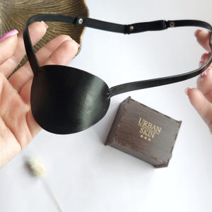 Puede incluir: Un parche para el ojo de cuero negro con una correa ajustable. La correa tiene varios broches para un ajuste personalizado. El parche para el ojo se muestra sobre un fondo blanco.