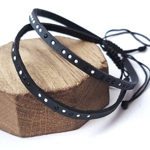 May include: A black leather bracelet featuring a series of small, evenly spaced white dots along the band. The bracelet has an adjustable sliding knot closure made from thin black cord, allowing for a flexible fit around the wrist or ankle.
