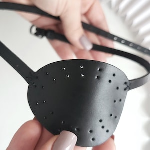 Può includere: Una benda sull'occhio nera con piccoli fori, tenuta da una persona. La benda sull'occhio è realizzata in un materiale liscio e ha un cinturino che va intorno alla testa. Il cinturino ha una fibbia per la regolazione.