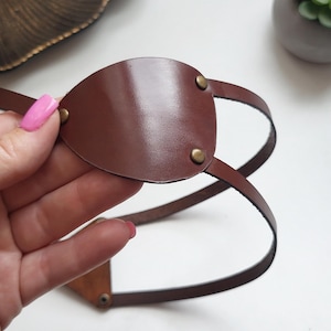 Può includere: Una benda per occhio in pelle marrone con un pezzo frontale liscio a forma di goccia, fissato con due piccoli rivetti metallici color ottone. La benda include sottili cinturini in pelle regolabili che si avvolgono per formare una fascia.