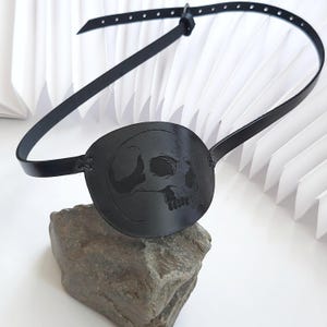 Peut inclure: Cache-œil en cuir noir avec un motif de crâne. Il est maintenu en place par une fine lanière réglable. L'accessoire est posé sur une pierre grise.