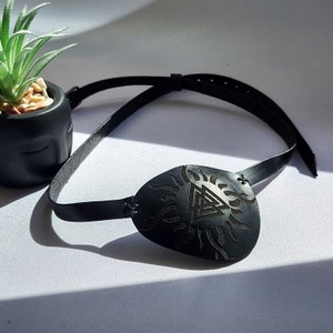 May include: A black leather eye patch with a black and white design of a sun with a triangle inside. The eye patch is attached to a black leather strap.