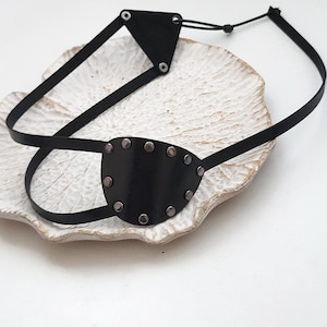 Pode incluir: Um tapa-olho preto com um escudo curvo e liso, adornado com rebites metálicos ao longo da borda. O tapa-olho está preso a um sistema de tiras pretas finas que inclui uma peça de conexão triangular e um cordão ajustável para fixação.