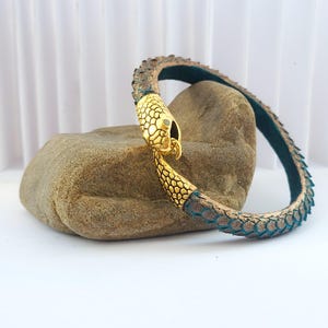 May include: A gold snake-shaped bracelet with a textured, green leather band. The bracelet is resting on a brown rock.