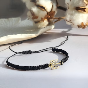 Op de afbeelding: Een zwart koordarmband met een gouden drietand bedel. De armband is verstelbaar en kan door mensen van alle maten worden gedragen.