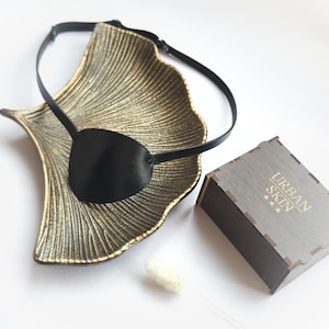 Könnte beinhalten: Ein schwarzes Leder-Augenpflaster mit verstellbarem Band, auf einem goldenen Dekotablett präsentiert. Das Augenpflaster ist in einer Holzkiste mit dem Aufdruck "URBAN SKIN" verpackt.
