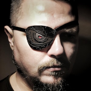 May include: A black eye patch with a detailed, mechanical design and a red accent. The eye patch is worn over one eye, with the frame of the glasses visible. The person has a beard and mustache.