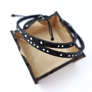 May include: Two black leather bracelets with white dots, displayed in a wooden box.