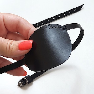 May include: A black leather eye patch featuring a smooth, rounded shield and a thin adjustable strap with a metal buckle. The word Chara is embossed in a cursive script on the upper right side of the patch, which is connected to the strap by stitching.