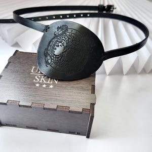 May include: Black leather eye patch with an embossed dragon design and a matching adjustable strap. The eye patch rests on a wooden box with the text "U SKIN" and three stars. The background is a white, folded paper fan.