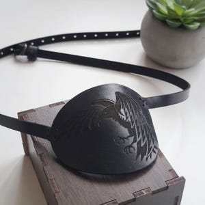 May include: A black leather eye patch featuring an embossed graphic of a raven in profile. The eye patch includes a thin, adjustable black leather strap with a metal buckle, designed to secure the piece comfortably around the head.