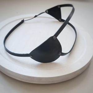 May include: A black leather eye patch with a strap that goes around the head. The eye patch is secured with metal rivets. The design includes a triangular piece above the eye patch and a thin strap that goes around the back of the head.