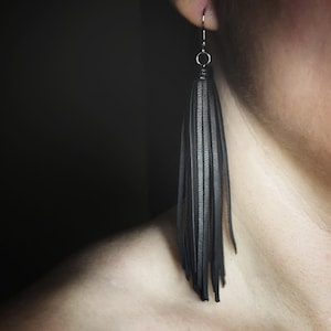 以下が含まれることがあります： 黒い革のタッセルイヤリング。イヤリングは銀色のフックから吊り下げられており、細長い黒革のストリップがフリンジ効果を生み出しています。イヤリングは暗い背景に対して表示されています。