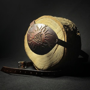 May include: A brown leather eye patch with a decorative metal design featuring a stylised sun with flames and a triangle symbol. The eye patch is on a wooden surface.