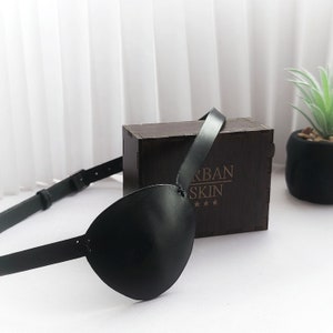 May include: A black leather eye patch with an adjustable strap. The strap is attached to the eye patch with a buckle. The eye patch is lying on a wooden box with the text "Urban Skin" engraved on it.