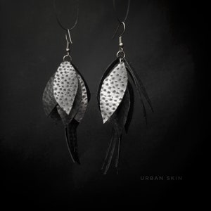 May include: A pair of black leather earrings with a layered leaf design. The earrings are hanging from silver hooks and have a textured surface. The text "URBAN SKIN" is visible at the bottom of the image.
