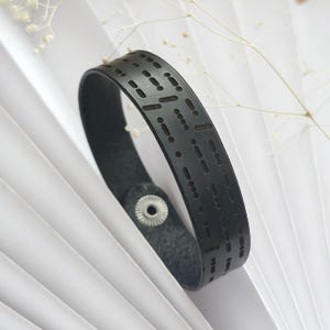 Może przedstawiać: Czarna skórzana bransoletka z wzorem kropek i kresek, typowy wzór dla brajla.
