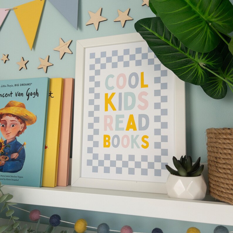 May include: A white framed print with a checkered background in grey and white. The text "COOL KIDS READ BOOKS" is written in colourful letters on the print. The letters are yellow, light blue, pink, and grey.