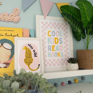 May include: Two framed prints with colourful text on a white shelf. The print on the left features a cheetah with the text "stay wild". The print on the right has a pink and white checkered background with the text "COOL KIDS READ BOOKS".