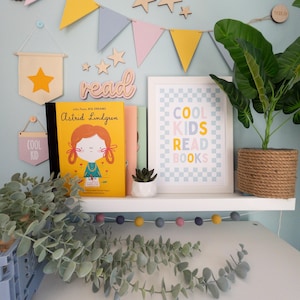 May include: A white shelf with a book titled "Little People, Big Dreams: Astrid Lindgren" and a framed print that says "Cool Kids Read Books" on a blue and white checkered background. There are wooden stars and flags hanging on the wall behind the shelf. A potted plant is on the shelf next to the book and print.