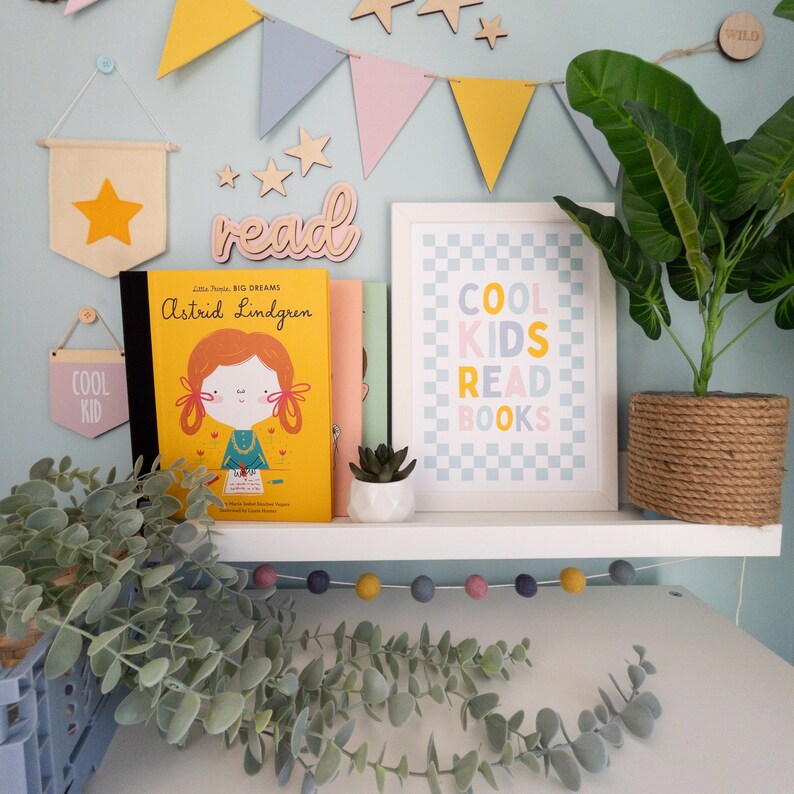 May include: A white shelf with a book titled "Little People, Big Dreams: Astrid Lindgren" and a framed print that says "Cool Kids Read Books" on a blue and white checkered background. There are wooden stars and flags hanging on the wall behind the shelf. A potted plant is on the shelf next to the book and print.