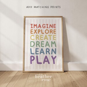 May include: A framed print featuring the words "IMAGINE, EXPLORE, CREATE, DREAM, LEARN, PLAY" in rainbow colours. The print is in a light brown wooden frame with a striped background. The text "ADD MATCHING PRINTS" is at the top, and "made by heather rose" is at the bottom.