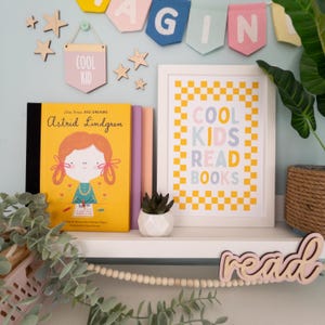 May include: A white shelf with a yellow and white chequered print framed poster that says "Cool Kids Read Books". There are two books on the shelf, one with a yellow cover and the text "Astrid Lindgren" and the other with a pink cover. There is a small wooden sign that says "Cool Kid" and a wooden sign that says "read" on the shelf.