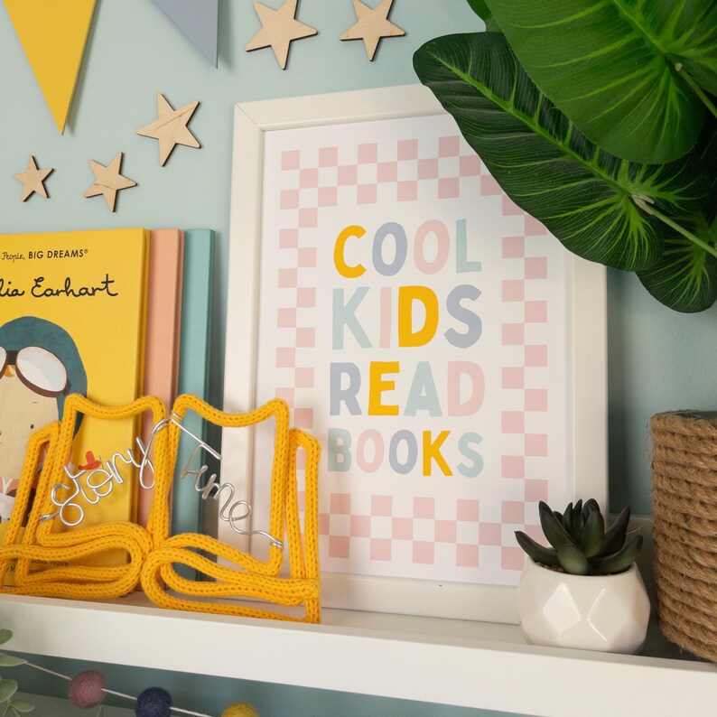 May include: A white framed print with a pink and white checkered background. The text "COOL KIDS READ BOOKS" is written in colourful letters on the print. The print is on a shelf with a yellow knitted bookend and other books.