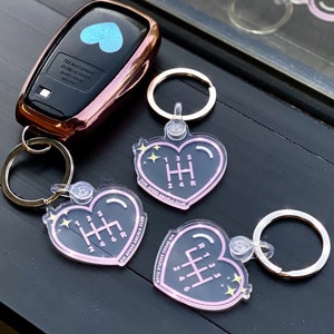 May include: A car key fob with a rose gold finish and a blue heart design, next to three heart-shaped keychains. The keychains feature a gear shift pattern and the text "FIVE SPEED ANGELS CLUB."