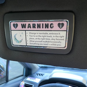 Puede incluir: Una visera de coche rosa y blanca con un cartel de advertencia que dice "WARNING" con un coraz&oacute;n a cada lado. El cartel tambi&eacute;n incluye una luna creciente con estrellas y el texto "Change is inevitable, embrace it. You're on the right track, in the right place, at the right time, stay focused. Allow yourself multidimensionally. Everything you need is within you."