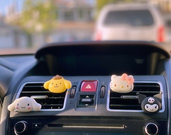 May include: Four colorful cartoon character air fresheners hanging from the air vents of a car's dashboard. The characters are a yellow dog, a white cat, a pink Hello Kitty, and a black and white character with a skull.